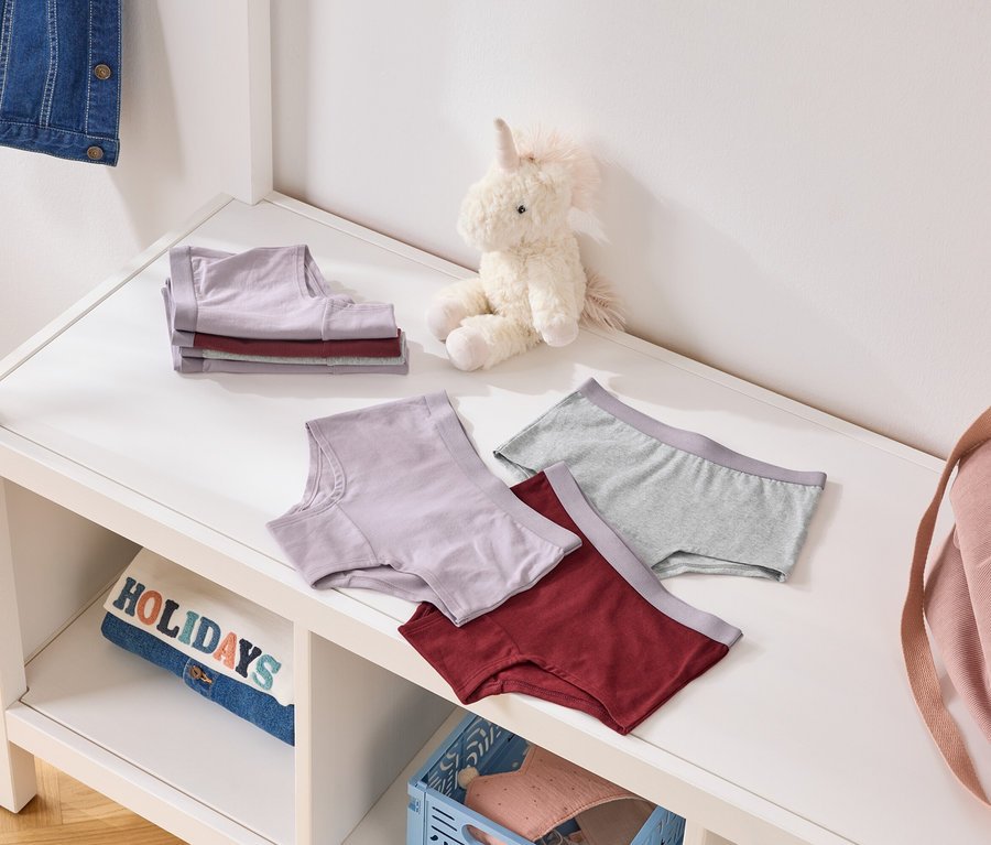 Sur une étagère, des culottes pliées et dépliées violettes, grises et bordeaux, à côté d'une licorne en peluche et d'un sac rose.