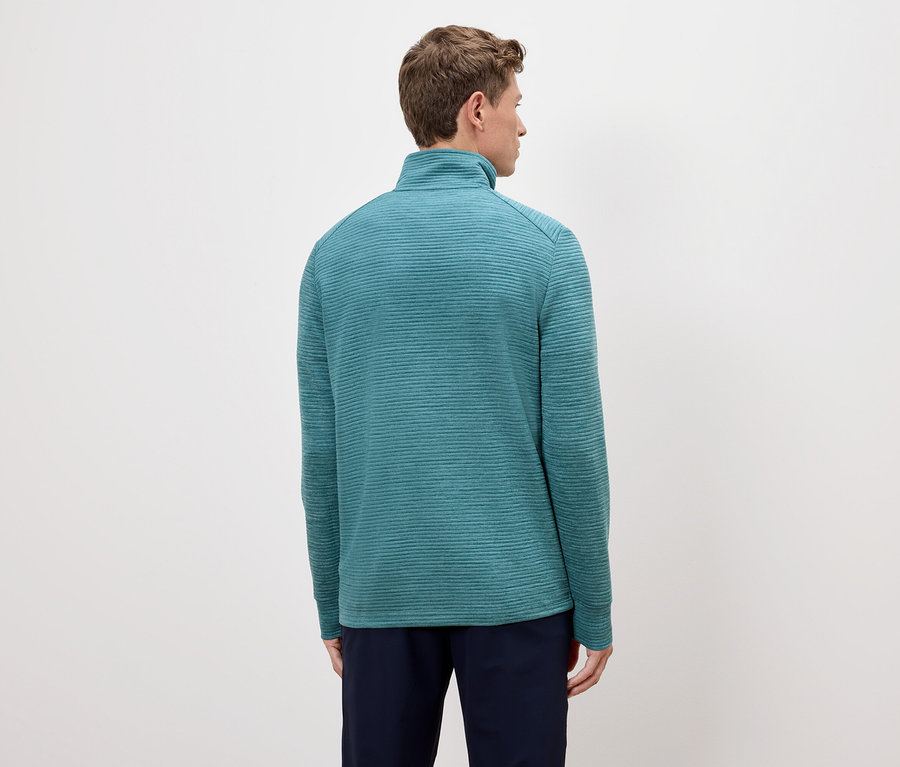 Vue arrière d'un homme aux cheveux châtain clair portant un pull turquoise zippé et un pantalon bleu marine sur un fond crème.