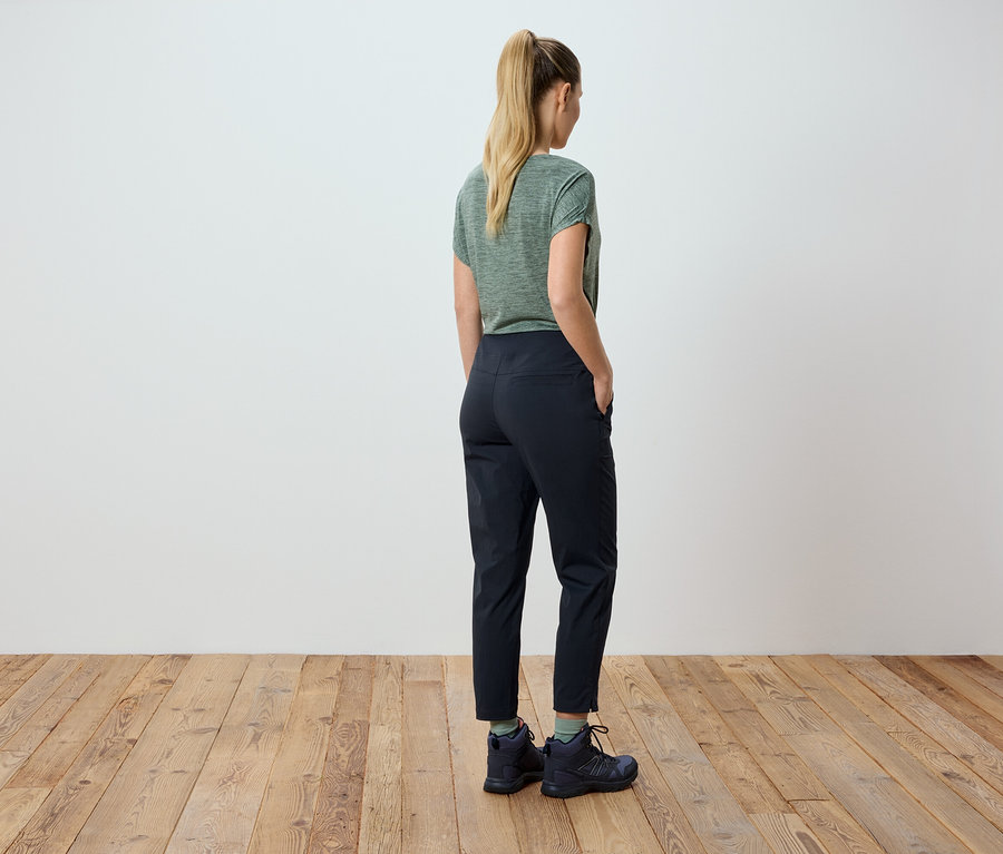 Femme blonde portant un t-shirt vert et un pantalon noir, les mains dans les poches.