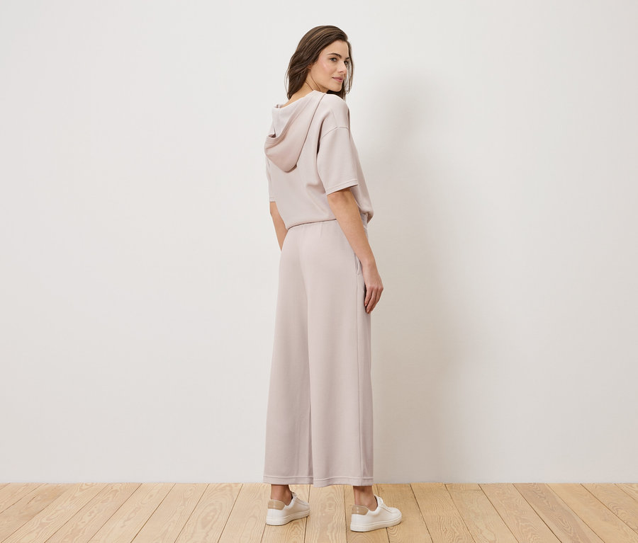 Une femme se tient debout dans un tailleur pantalon rose clair et des baskets blanches, la tête tournée par-dessus son épaule.