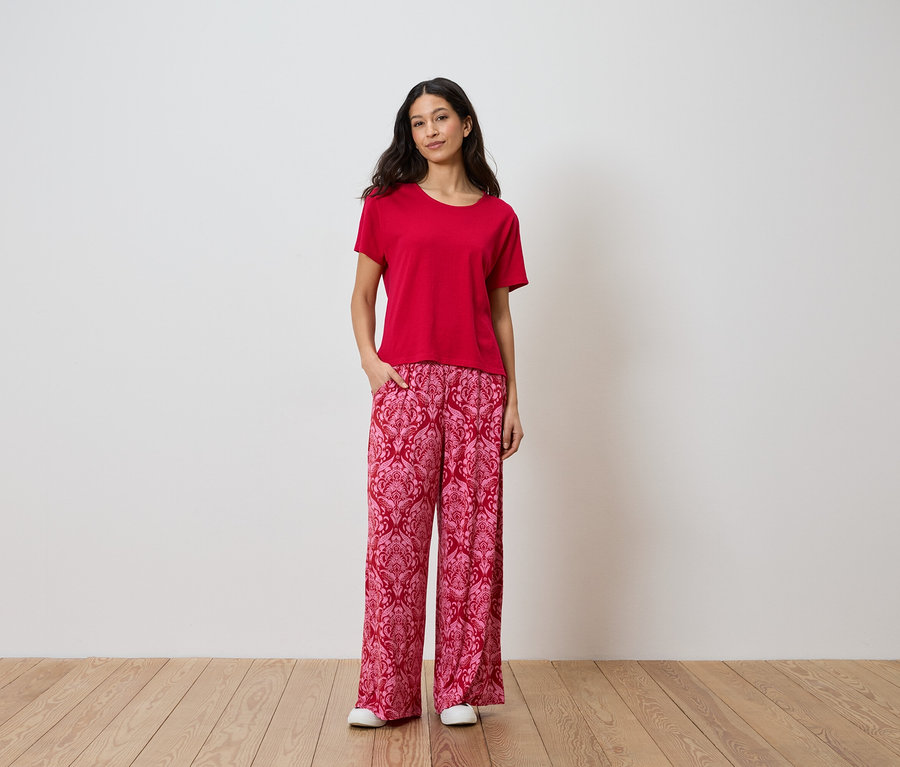 Femme portant un t-shirt rouge et un pantalon à motifs devant un mur blanc.