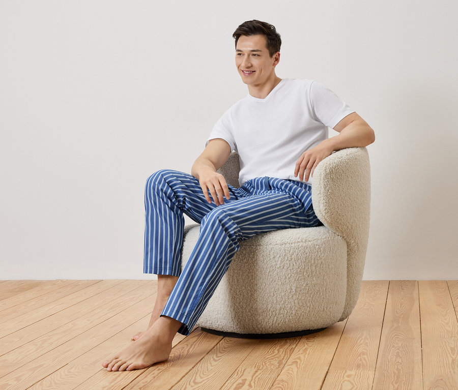Un homme est assis pieds nus dans un fauteuil beige. Il porte un t-shirt blanc et un pantalon de pyjama rayé bleu et blanc.