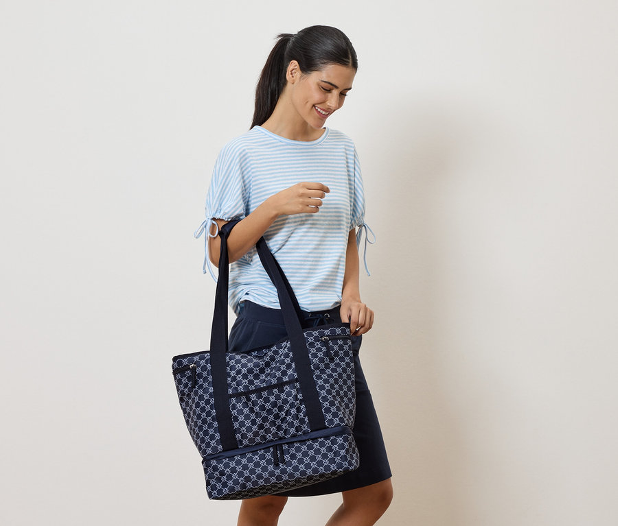 Une femme souriante portant un t-shirt rayé bleu et blanc et une jupe bleu marine tient un sac fourre-tout bleu marine à motifs.