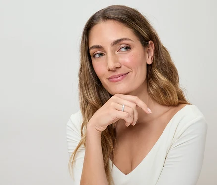 Femme aux longs cheveux ondulés et haut blanc, portant une bague argentée au doigt, regardant pensivement sur le côté.