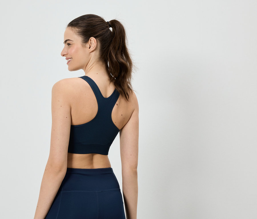 Une femme avec une queue de cheval, vue de dos, porte un soutien-gorge de sport bleu marine et un legging sur fond blanc.