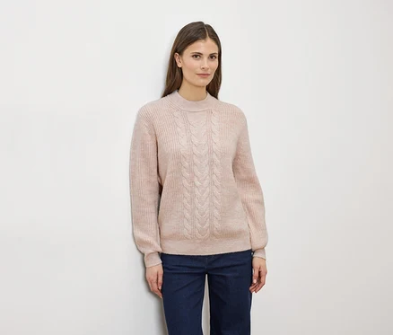 Une femme aux longs cheveux bruns porte un pull en maille torsadée rose clair et un jean bleu devant un mur blanc.