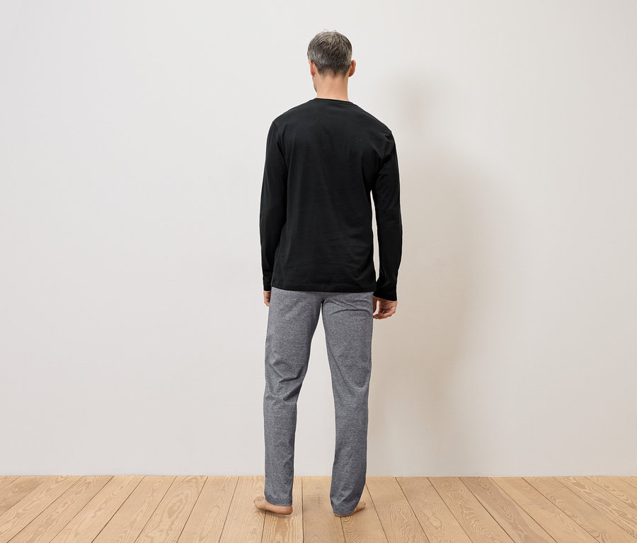 Homme vu de dos portant un t-shirt noir à manches longues et un pantalon gris devant un mur sur un plancher en bois.