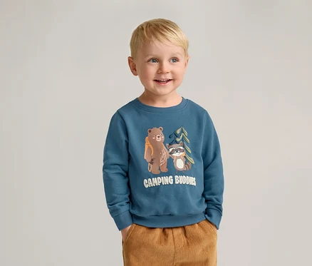 Un garçon blond souriant porte un sweat-shirt bleu avec un motif d'ours et de raton laveur et un pantalon marron.