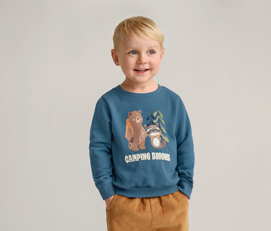 Un garçon blond souriant porte un sweat-shirt bleu avec un motif d'ours et de raton laveur et un pantalon marron.