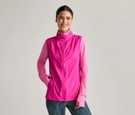 Une femme souriante porte un gilet rose sur un haut rose à manches longues et un legging rayé.