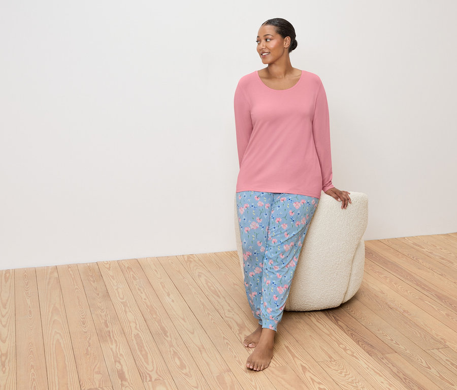 Femme portant un haut rose et un pantalon bleu à fleurs, appuyée sur un pouf beige.