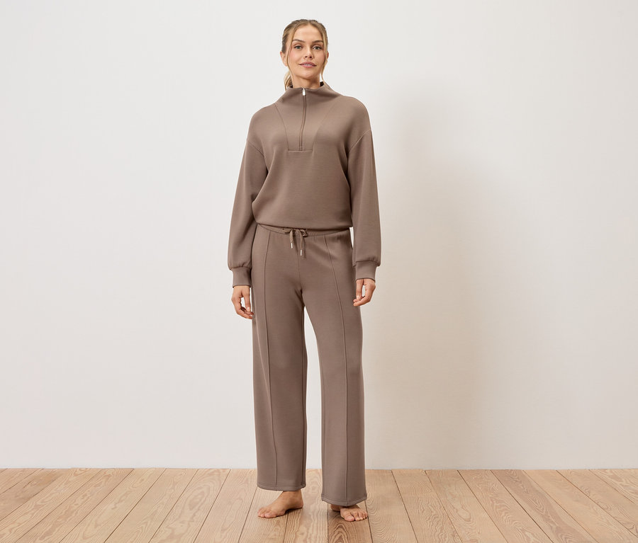 Une femme se tient pieds nus sur un plancher en bois et porte un survêtement marron.