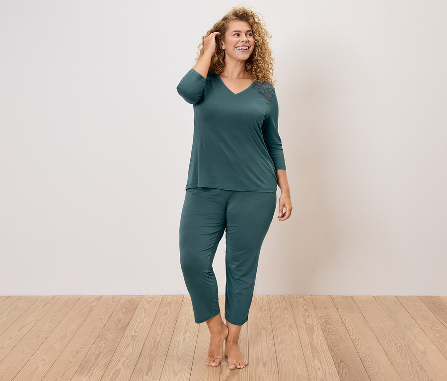 Femme aux cheveux bouclés portant un ensemble de pyjama vert foncé composé d'un haut à col en V et d'une application de dentelle sur l'épaule et d'un pantalon.