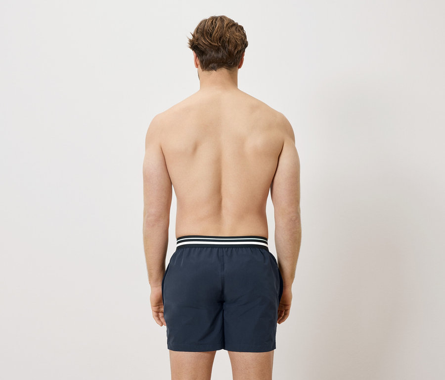 Vue arrière d'un homme torse nu portant un short de bain bleu marine avec une bande blanche.