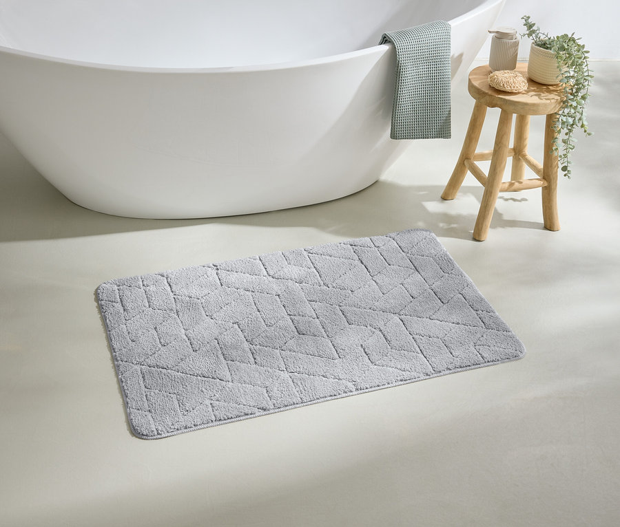 Un tapis de bain gris à motifs géométriques est posé devant une baignoire blanche. Une serviette verte est accrochée au bord de la baignoire.