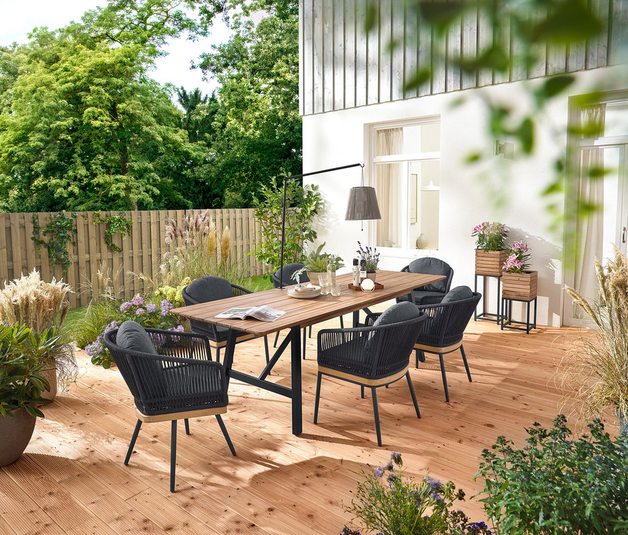 Sur une terrasse en bois se trouvent une table de jardin à rallonges avec structure rustique d'environ 200 à 250 cm, des chaises de jardin pivotantes avec tressage textile et des chaises de jardin avec tressage textile. On voit aussi 2 colonnes à plantes « Tinus » et une lanterne solaire à accu avec support de table.