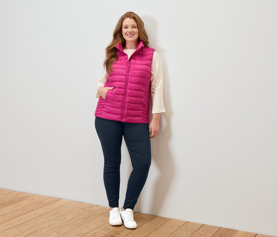 Femme portant une veste matelassée rose, un chemisier beige, un jean bleu et des baskets blanches, debout contre un mur blanc.