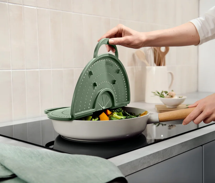 Une main soulève un couvercle-passoire vert d'une poêle avec des légumes sur une cuisinière.