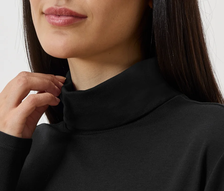Une femme aux cheveux foncés portant un pull à col roulé noir touche le col.