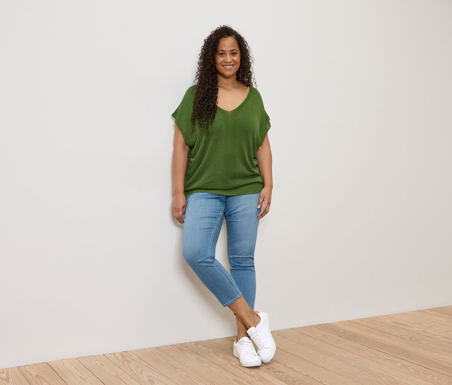 Une femme aux cheveux bouclés, portant un haut vert, un jean et des chaussures blanches, est appuyée contre un mur blanc.