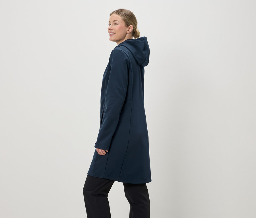 Une femme avec une queue de cheval porte un manteau softshell bleu marine à capuche et un pantalon noir sur un fond clair.