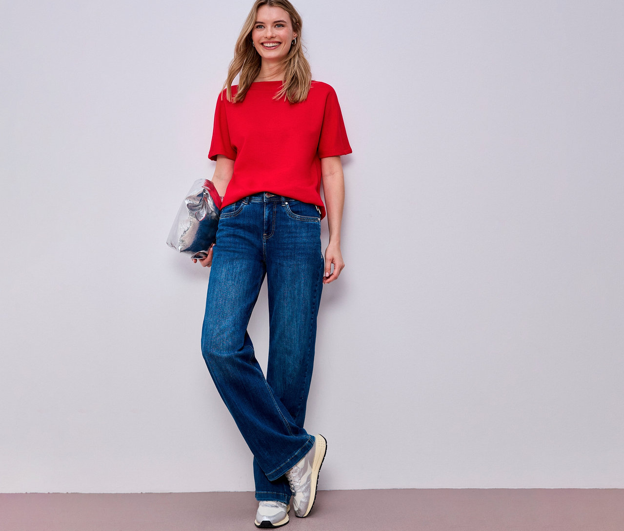 Femme portant une chemise à manches courtes Street One rouge, un jean bleu et des baskets argentées tenant un sac argenté.