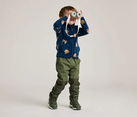 Un garçon portant une veste en polaire bleu marine à motif d'ours regarde à travers des jumelles jouets. Il porte un pantalon et des bottes verts.