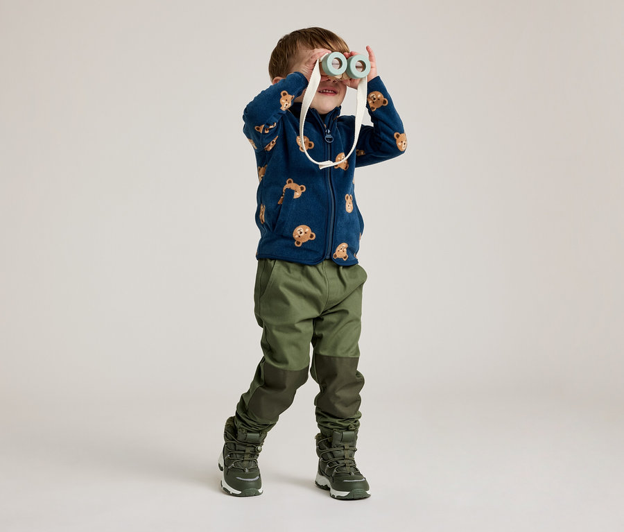Un garçon portant une veste en polaire bleu marine à motif d'ours regarde à travers des jumelles jouets. Il porte un pantalon et des bottes verts.