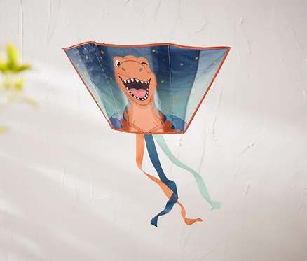 Un cerf-volant trapézoïdal avec un dinosaure orange souriant et des étoiles est accroché à un mur blanc texturé. Des rubans orange, bleu foncé et bleu clair pendent en bas.