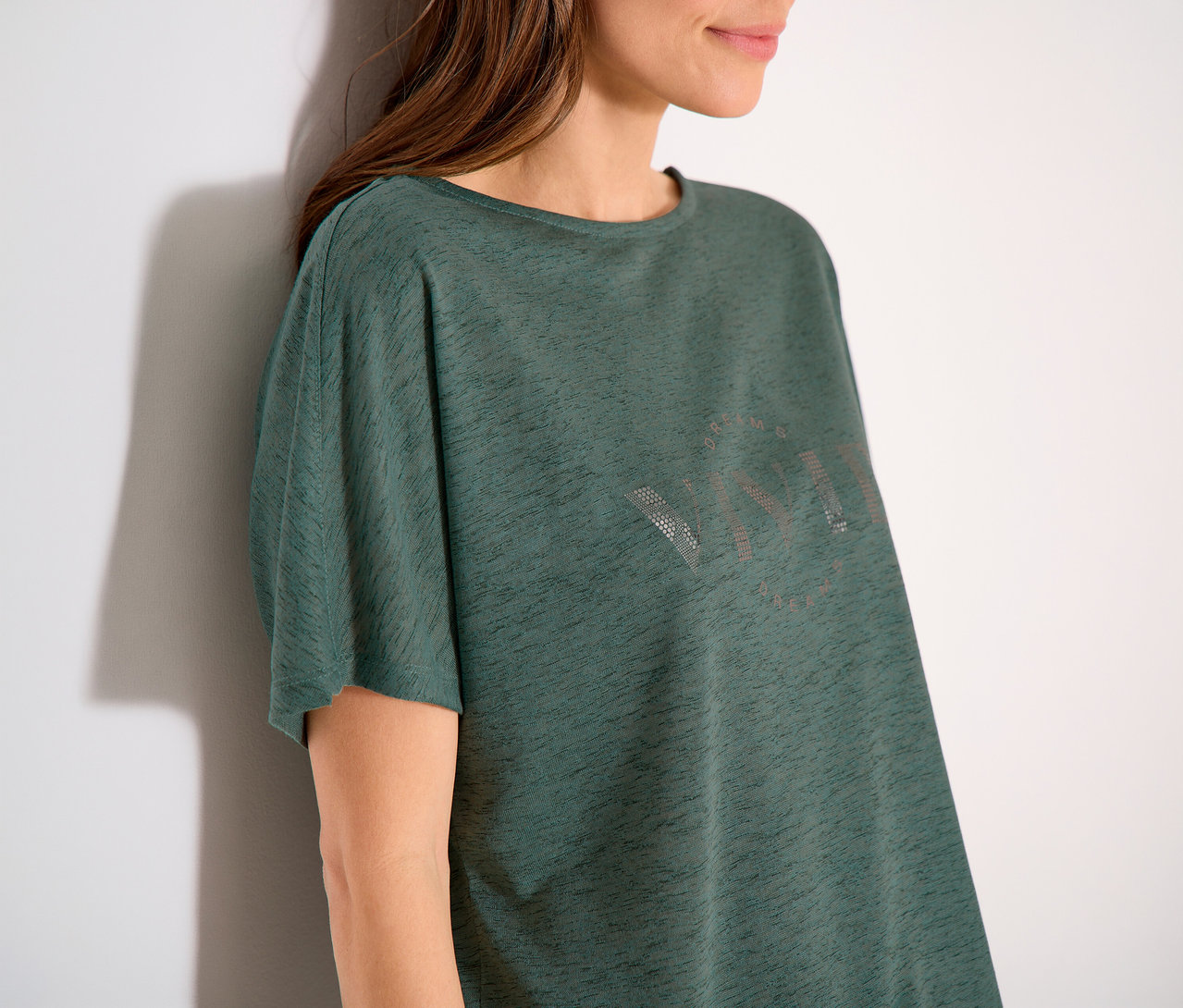 Femme appuyée contre un mur portant un t-shirt vert avec l'inscription VIVID.
