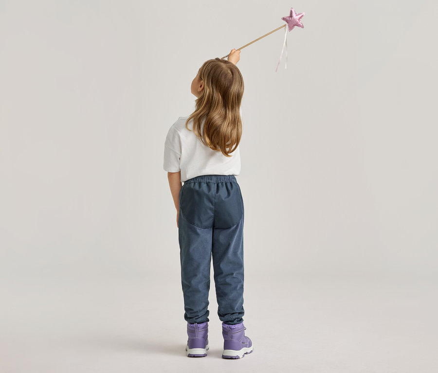 Vue de dos d'une fillette blonde tenant une baguette magique avec une étoile rose. Elle porte un pantalon bleu et des bottes violettes.