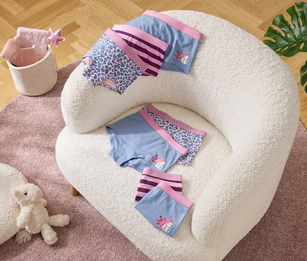 Sur un fauteuil sont posés des slips bleus avec des bordures violettes et des motifs. À côté, un panier avec des textiles et un ours en peluche sur un tapis.