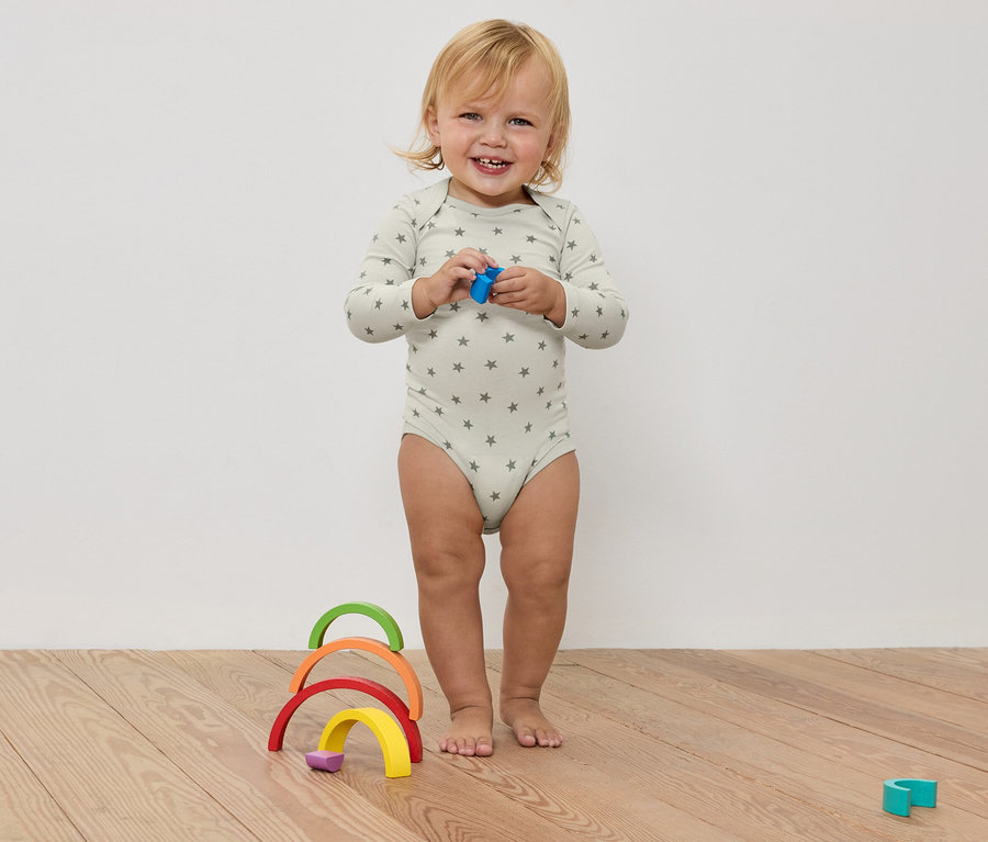 Un bébé souriant en body étoilé tient un jouet dans ses mains, devant un arc-en-ciel en bois coloré sur un plancher en bois.