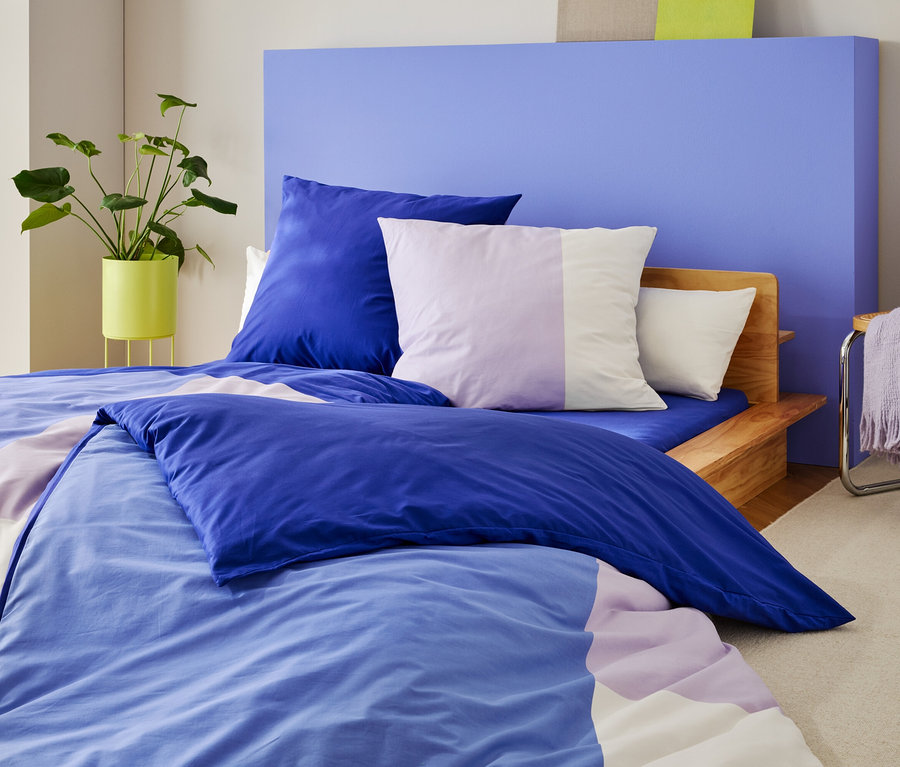 Un lit avec des draps et des coussins bleu-violet se dresse devant un mur bleu, à côté d'une plante et d'une chaise avec une couverture.