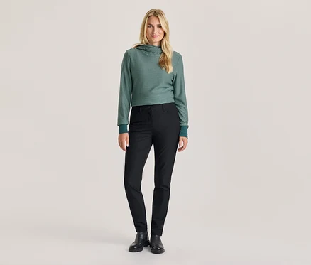 Une femme se tient debout, vêtue d'un sweat à capuche rayé vert et blanc, d'un pantalon noir et de bottes noires.