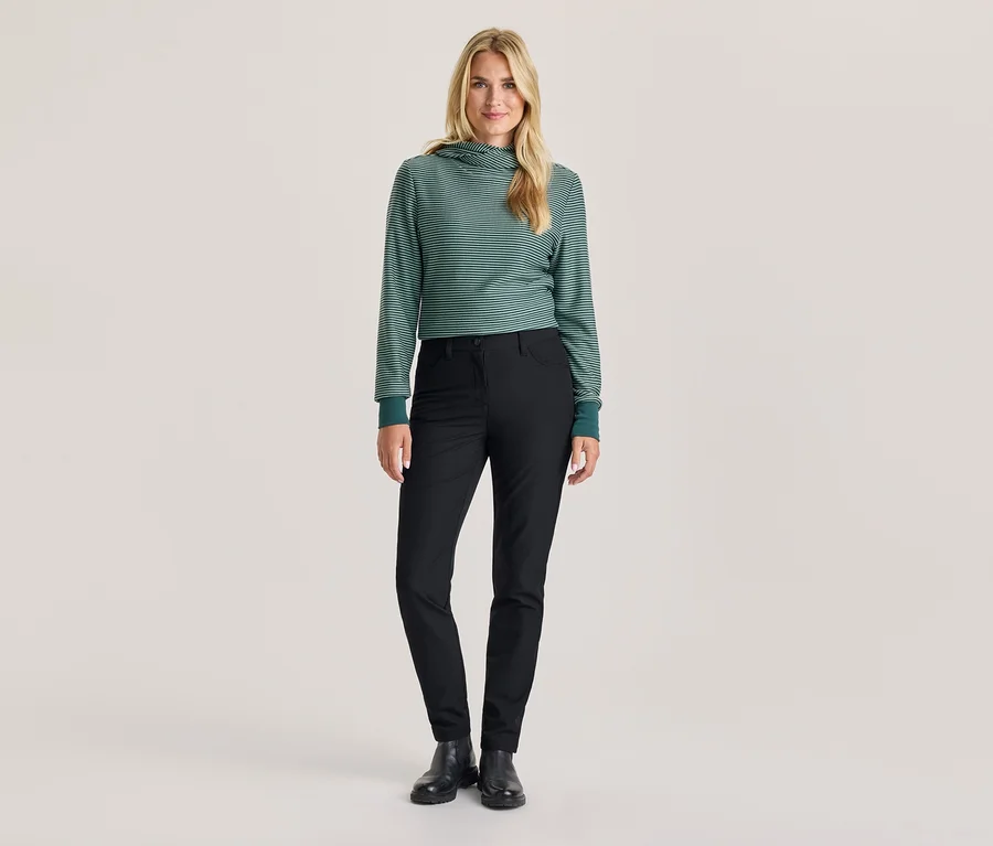 Une femme se tient debout, vêtue d'un sweat à capuche rayé vert et blanc, d'un pantalon noir et de bottes noires.