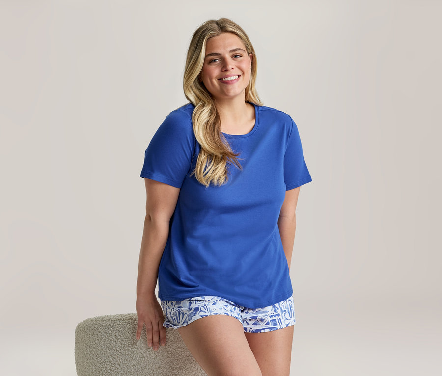 Femme souriante, vêtue d'un pyjama shorty bleu roi avec un haut uni et un short à motifs, appuyée sur un tabouret.