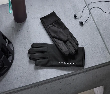 Casque de vélo noir, gourde foncée, gants noirs avec points réfléchissants et écouteurs noirs posés sur une surface en béton gris.