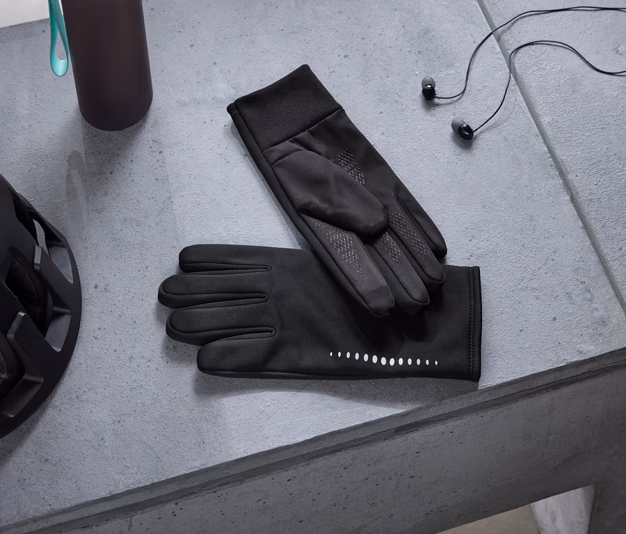 Casque de vélo noir, gourde foncée, gants noirs avec points réfléchissants et écouteurs noirs posés sur une surface en béton gris.