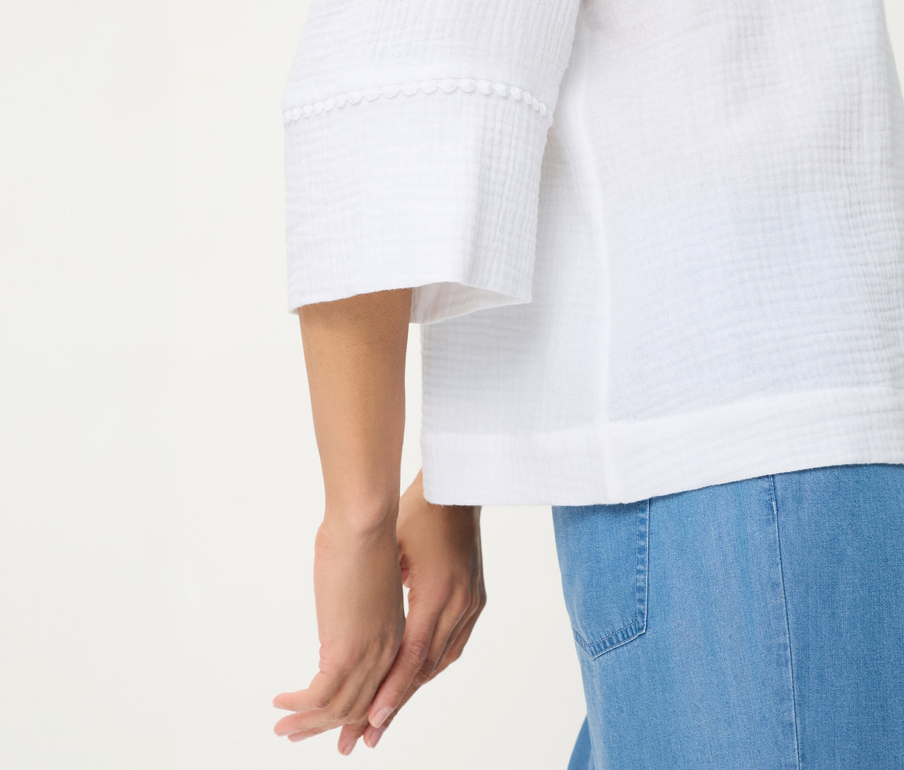 Gros plan d'une personne portant une blouse tissée Olsen blanche avec un tissu texturé et une bordure décorative sur les manches, associée à un jean bleu clair. Les mains de la personne sont jointes.