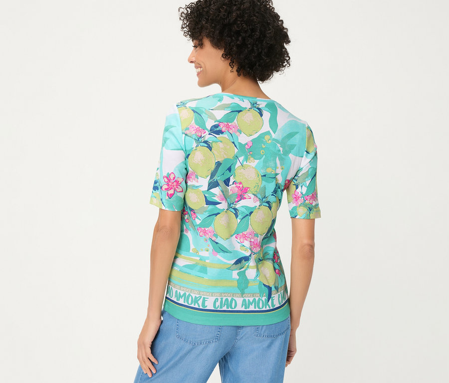 Femme de dos portant un T-shirt Olsen à motif citrons, feuilles et fleurs roses, avec une bordure rayée et l'inscription « AMORE CIAO », et un jean bleu.