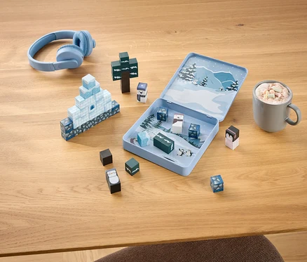 Des écouteurs bleus, un jeu de construction hivernal ouvert avec des blocs formant un igloo et un arbre, et une tasse de chocolat chaud avec des guimauves sur une table en bois.