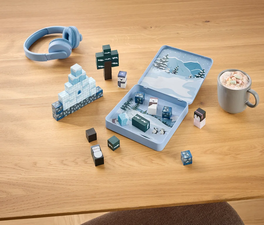 Des écouteurs bleus, un jeu de construction hivernal ouvert avec des blocs formant un igloo et un arbre, et une tasse de chocolat chaud avec des guimauves sur une table en bois.