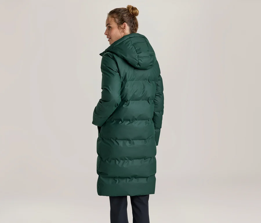 Femme vue de dos, légèrement tournée vers la droite, portant un long manteau matelassé vert foncé avec une capuche. Ses cheveux sont attachés en chignon.