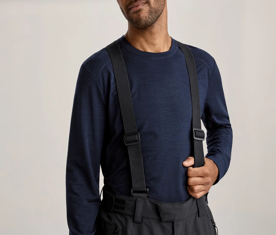 Homme portant un t-shirt à manches longues bleu foncé et un pantalon noir avec des bretelles, tenant une bretelle de sa main droite.