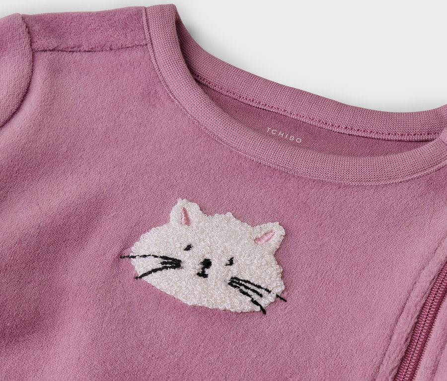 Gros plan sur une grenouillère bébé en polaire rose avec une broderie de tête de chat blanche et duveteuse, des moustaches noires et des oreilles roses. Une fermeture éclair diagonale et des boutons-pression à l'épaule sont visibles, 'TCHIBO' est imprimé sur le col.