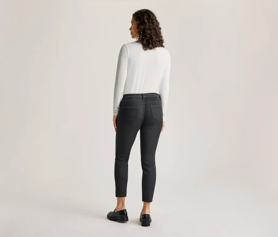 Une femme est debout, dos au spectateur, regardant légèrement vers sa gauche. Elle porte un haut blanc à manches longues, un tregging gris foncé – coupe « Nelly » et des mocassins noirs.