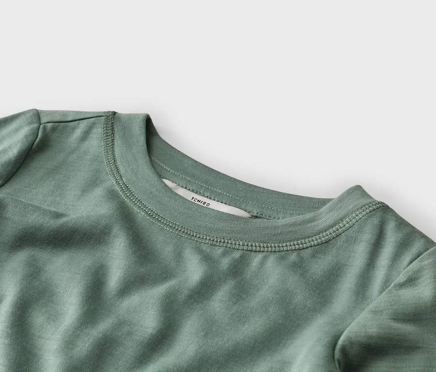 Gros plan d'un t-shirt vert sauge, montrant le col rond côtelé avec des coutures visibles et une étiquette blanche "TCHIBO" à l'arrière du cou.