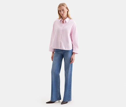 Femme debout, portant un chemisier oversize Seidensticker rose à rayures et un jean bleu.