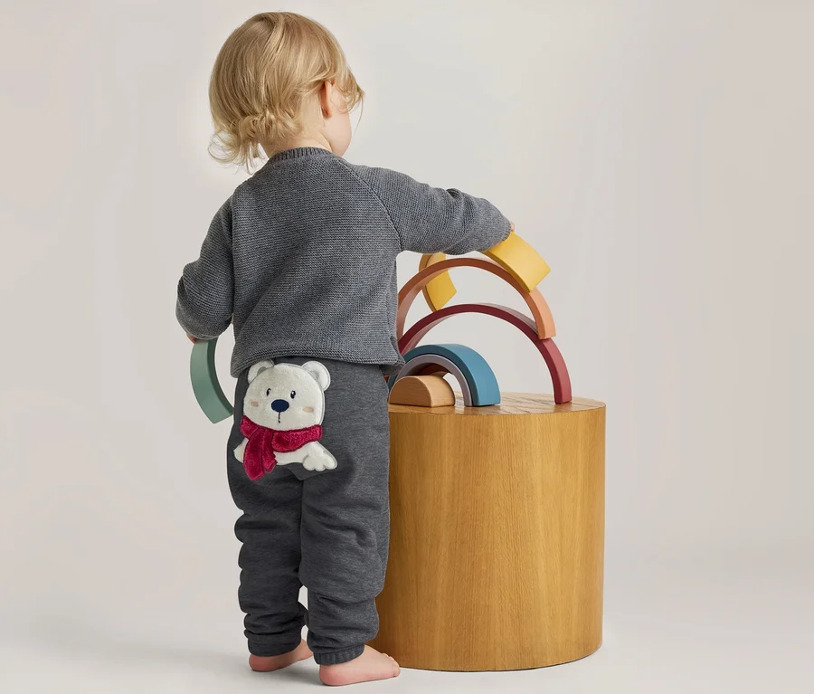 Enfant en bas âge de dos, en pull et pantalon gris avec un ours polaire appliqué, jouant avec des jouets d'empilement arc-en-ciel en bois sur un tabouret.